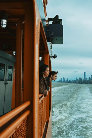 Two people lean over the railing of a ferry boat gazing at a city skyline across the water on a cloudy day.