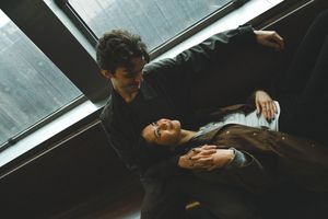 A man and woman in dark clothing share an intimate moment, with the woman lying across the man's lap indoors.
