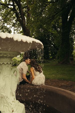 Couple sharing a romantic kiss beside a white ornate fountain in a lush green park setting.