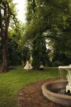 Bride in white dress walks toward groom near ornate fountain in lush green garden park setting.