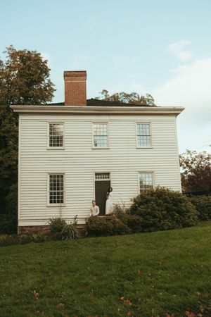 Historic white clapboard house with multiple windows sits on a grassy hillside under blue skies.