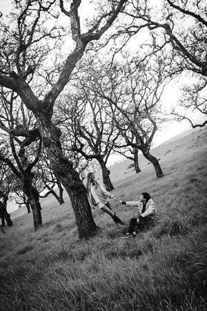 Black and white photo of a couple holding hands among bare oak trees on a grassy hillside.