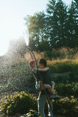 Four sequential shots showing someone playfully spraying water with a garden hose in a sunny backyard with sunflowers.