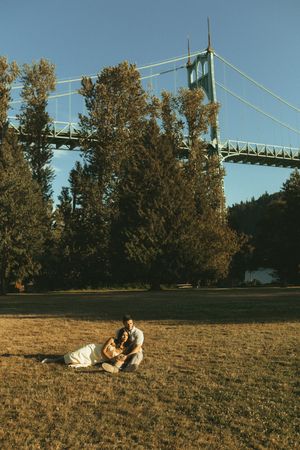Two people lounge on a grassy hill with autumn trees and a bridge visible in the background.