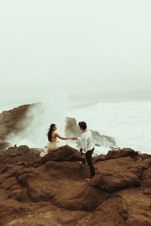 Two people stand on rocky coastal cliffs as ocean waves crash and spray dramatically against the shore.