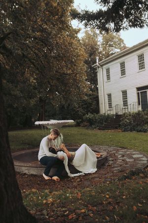 Two people lying on a stone patio in autumn with fallen leaves scattered around a white colonial house.