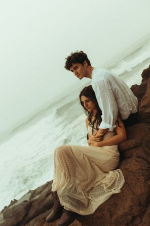 A person in a white dress sits contemplatively on rocky terrain against a misty white backdrop.