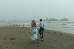 People walking along a misty beach shoreline with waves crashing in the distance on a cloudy day.