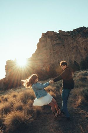 Couple dancing and holding hands at golden hour with sunburst and rocky cliffs in a scenic outdoor landscape.