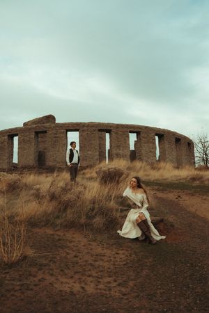 A figure in a flowing white dress poses near abandoned brick ruins on a barren hillside at dusk.