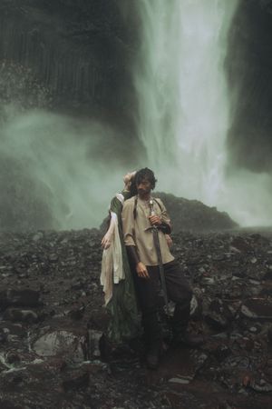 A person in a poncho stands on dark rocky terrain near a misty waterfall in moody atmospheric conditions.