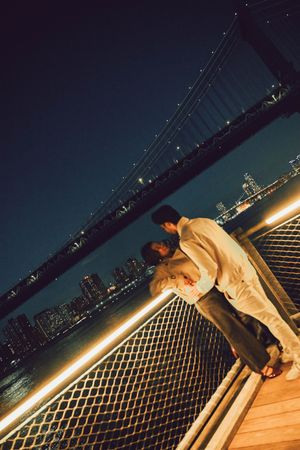 Couple embracing romantically on a waterfront deck at night with Manhattan Bridge and NYC skyline glowing behind them.