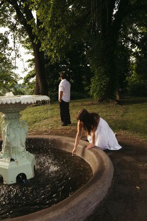 Woman in white dress touching water in ornate fountain while man in white shirt stands nearby in lush green park.