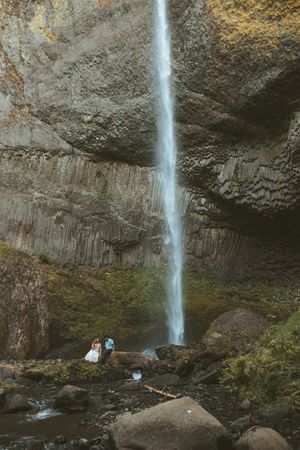 A tall waterfall cascades down a rocky cliff face with visitors viewing from a scenic overlook in the distance.