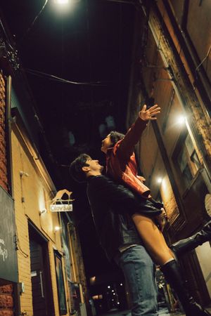 Person performing against a brick wall backdrop at night with overhead lighting creating dramatic shadows.
