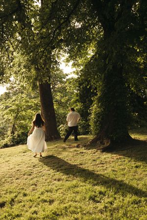 Couple walking toward sunlit trees in a lush green park, woman in white dress, man in dark pants.