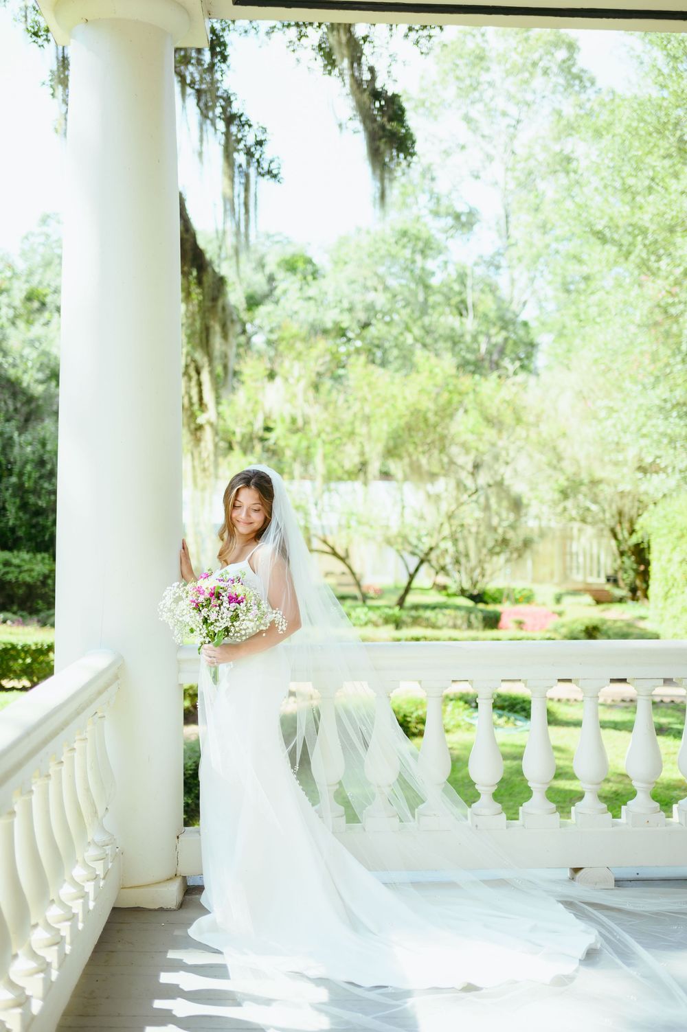 A bride in a flowing white gown holds a pink bouquet while standing on an elegant Southern plantation-style porch.