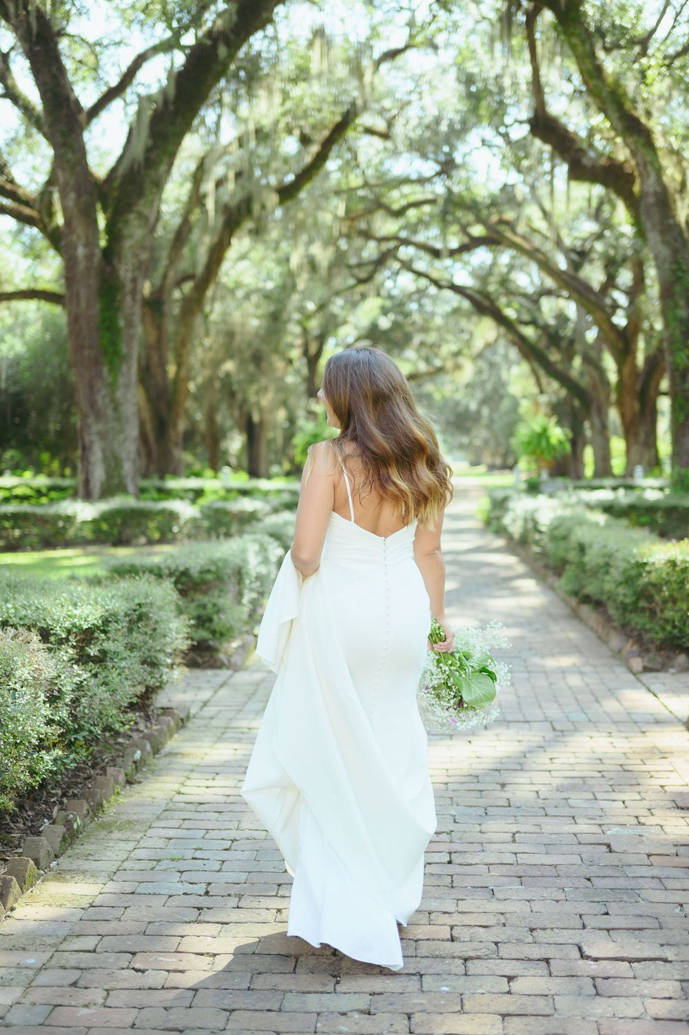 A bride in a flowing white dress walks down a brick path beneath Spanish moss-draped oak trees in a garden setting.