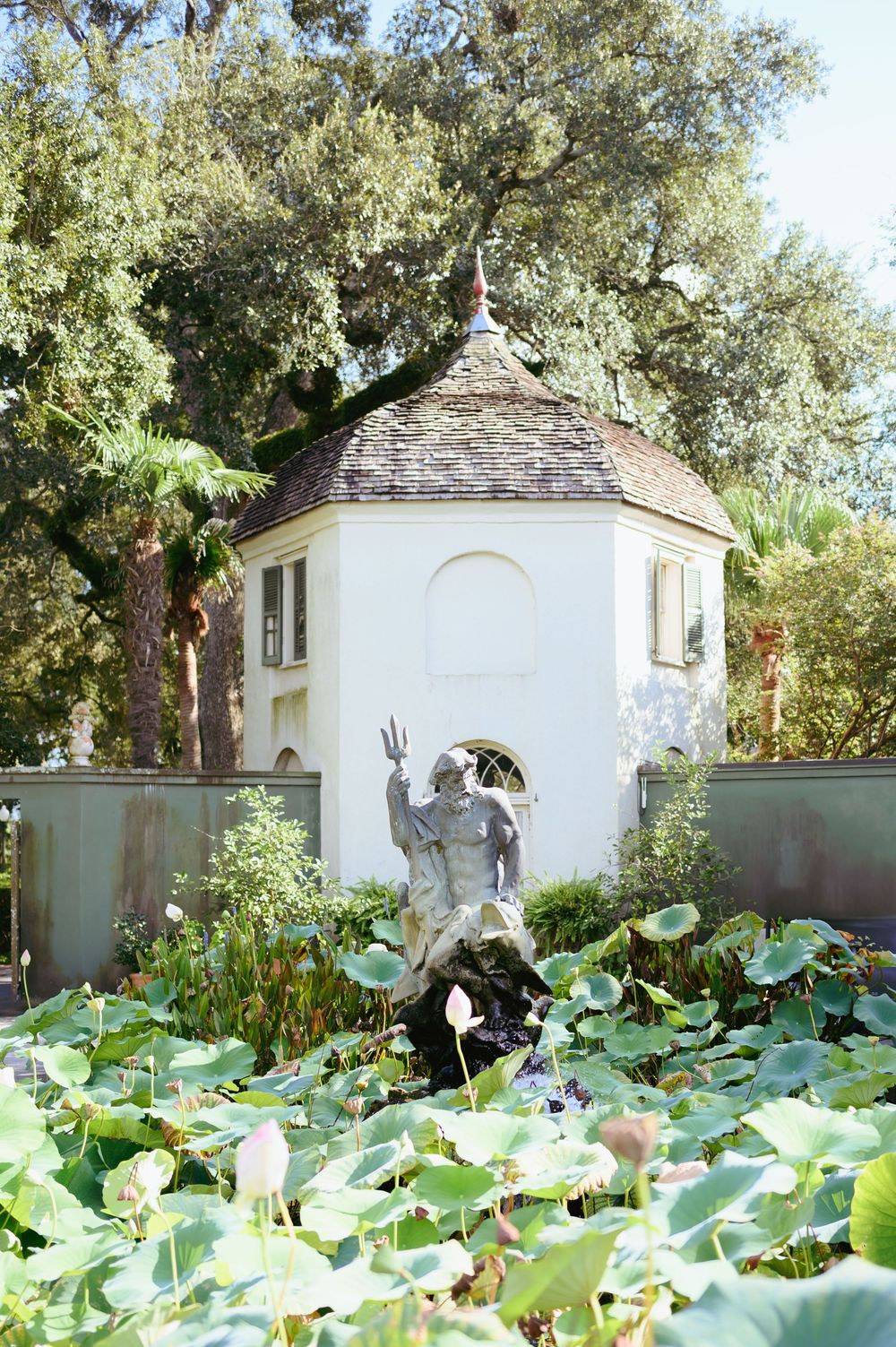 A white garden folly with a stone sculpture fountain stands among lush lotus leaves in a tranquil garden setting.