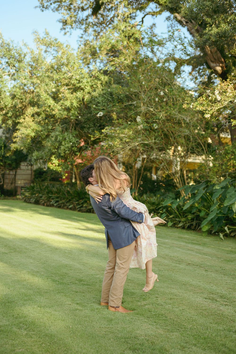 A couple embraces on a lush green lawn surrounded by mature trees on a sunny day.
