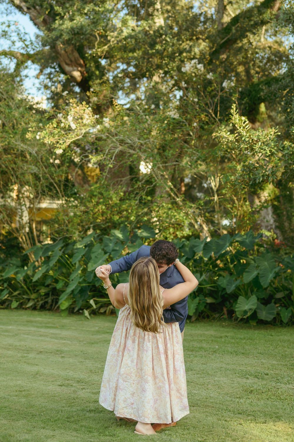 A couple shares an intimate dance moment together on a lush green lawn surrounded by tropical foliage.