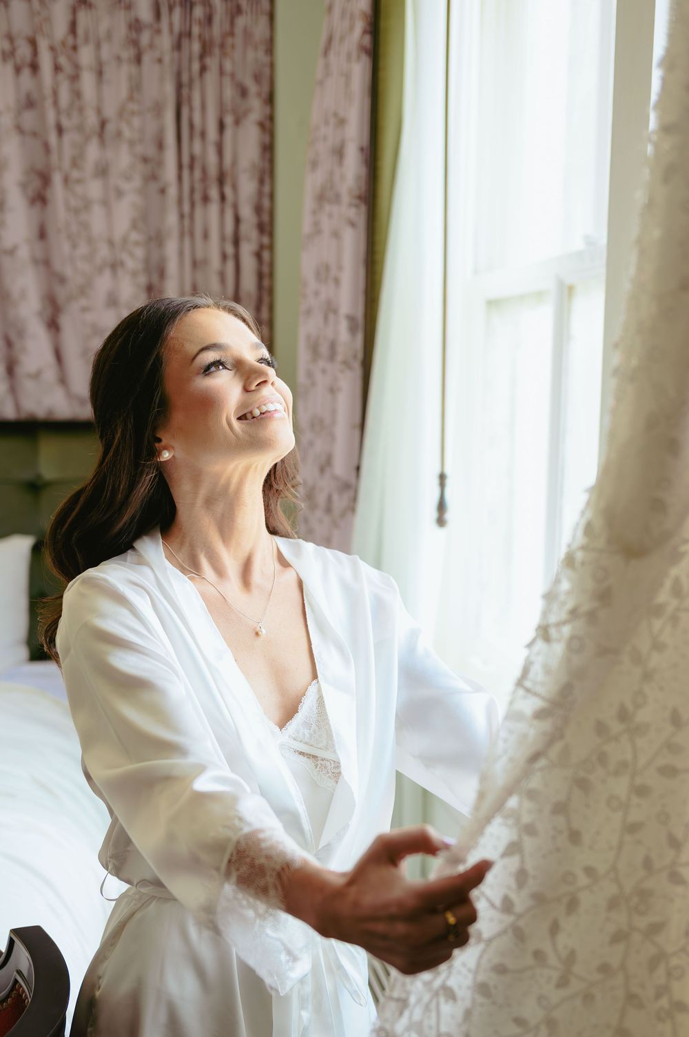 A person in a white robe smiles while opening curtains by a bright window.