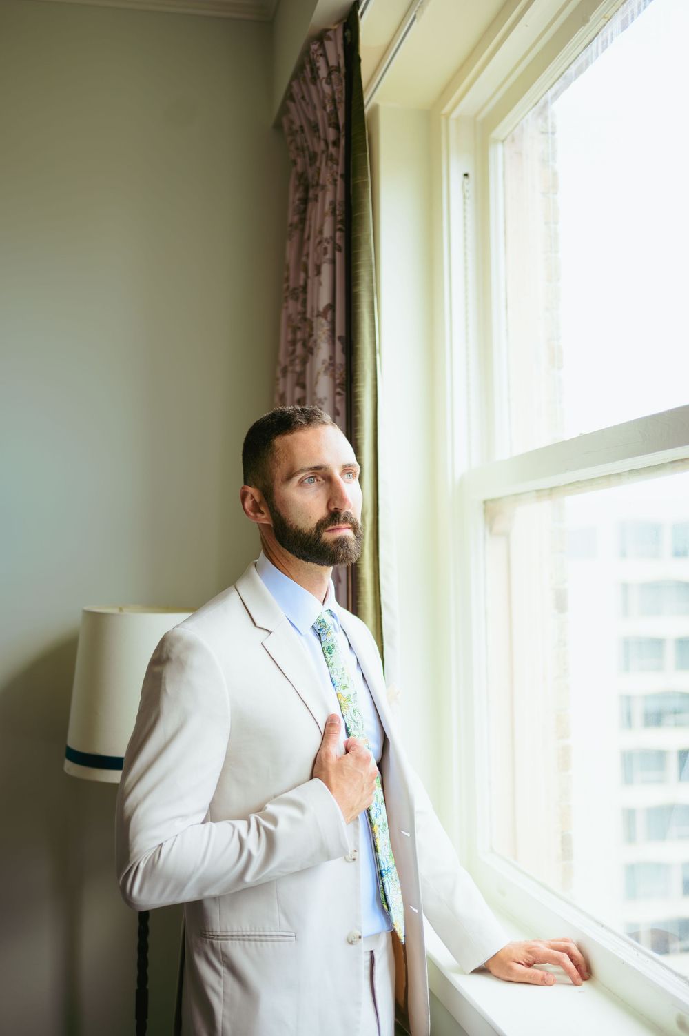 A person in a light gray suit and mint green tie stands thoughtfully by a bright window with curtains in a neutral room.