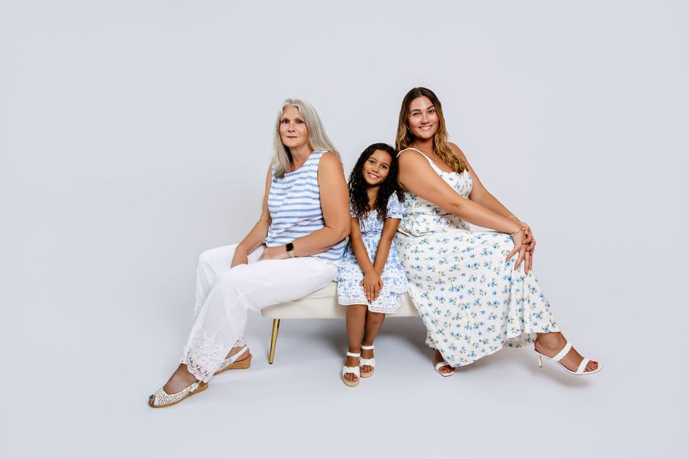 Three people in summer dresses sitting together on white bench against light background.