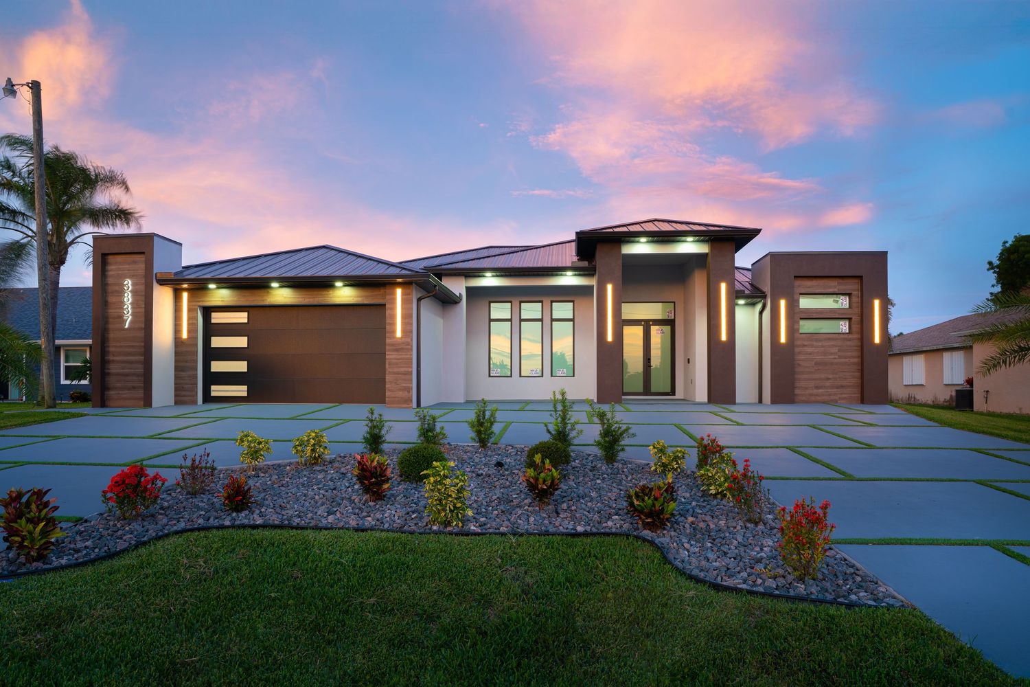 Contemporary gray house exterior with paver driveway and tropical landscaping.