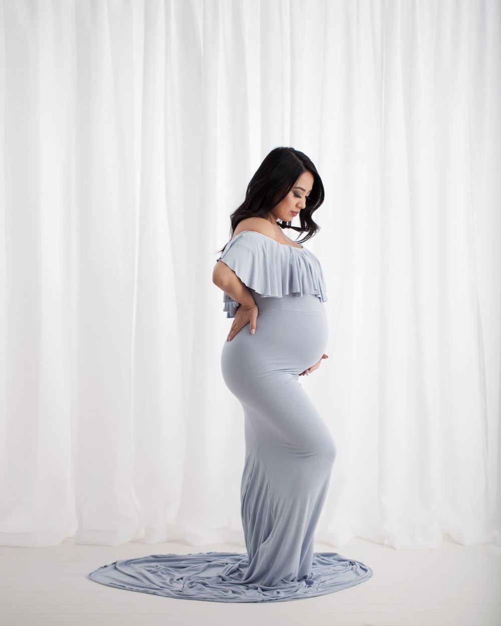 Artistic maternity portrait featuring gray floor-length dress against white backdrop.