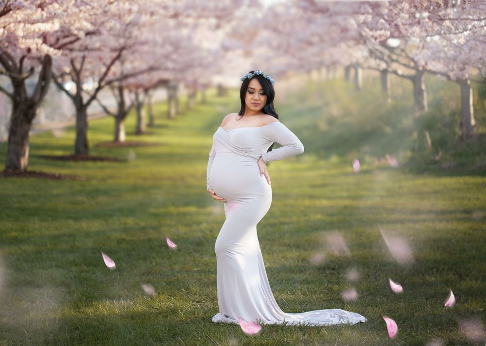 Expectant mother in flowing white gown poses among blooming cherry blossom trees in a sunlit garden.