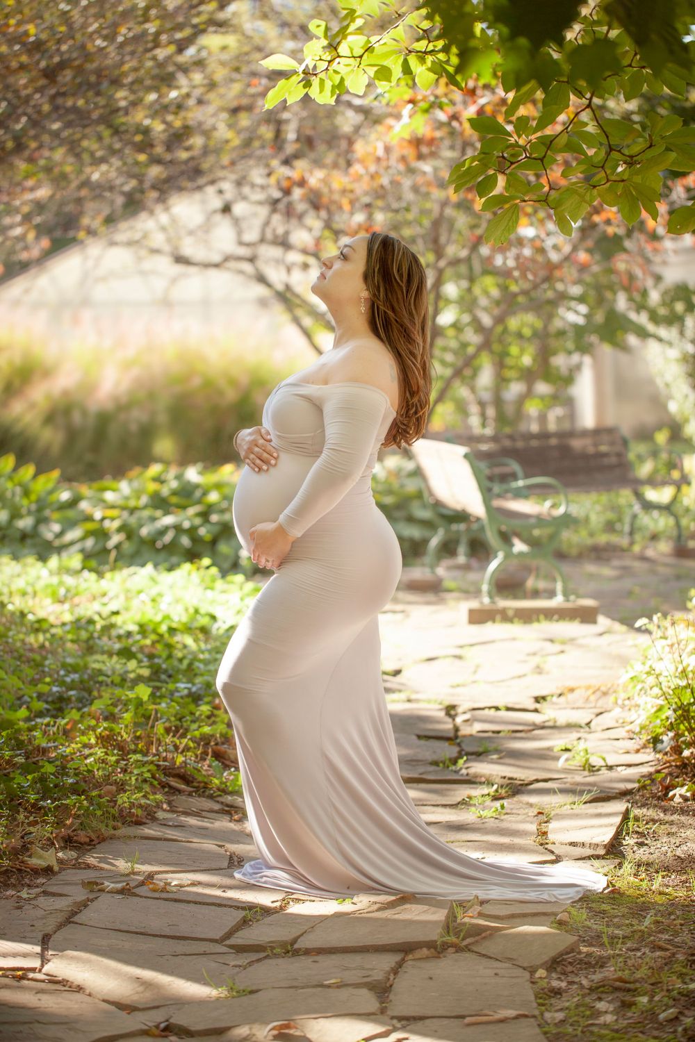 Dreamy maternity portrait on stone path with sunlit garden backdrop.