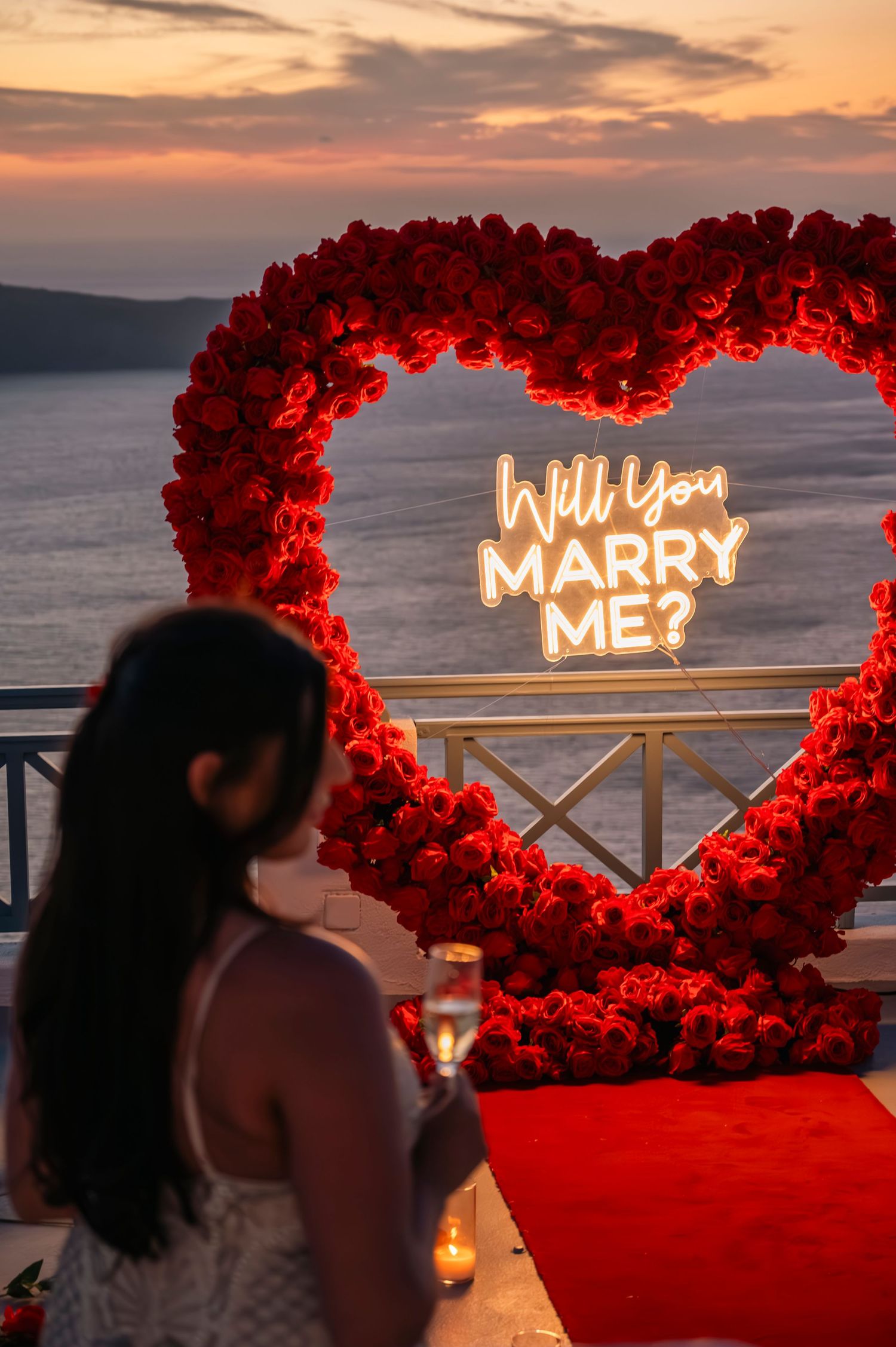Red rose heart arch with illuminated 'Will you marry me?' sign overlooking the sea at sunset.