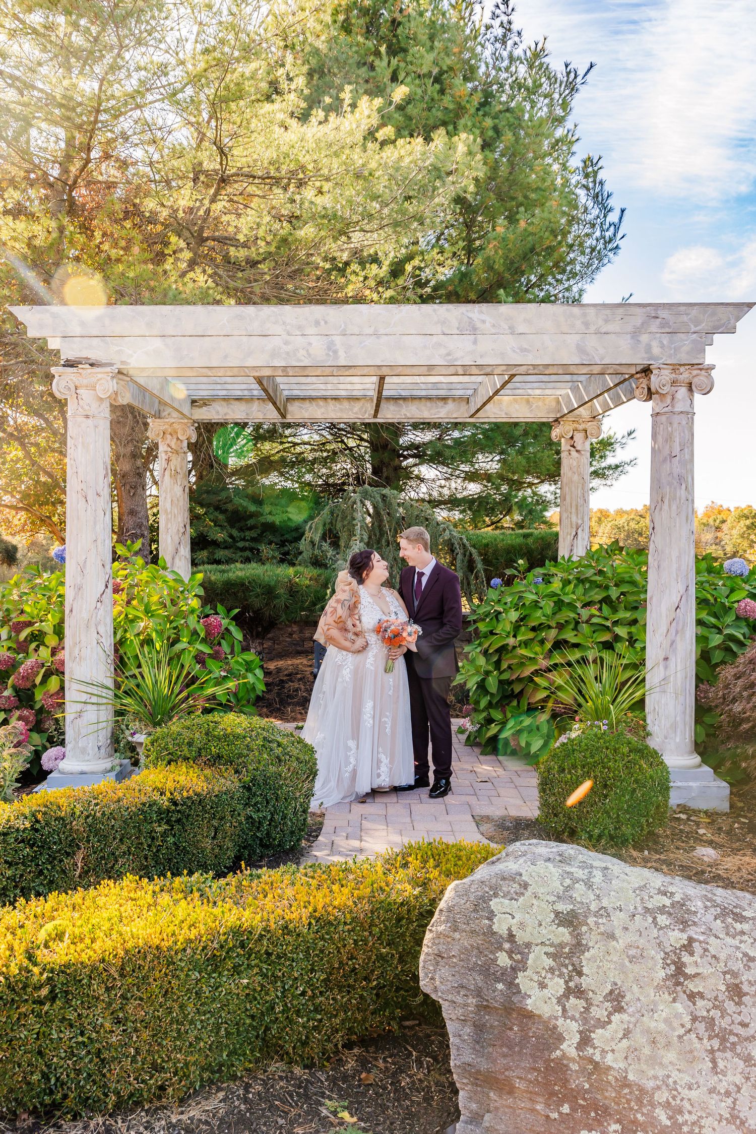 Wedding couple stands under elegant white pergola in a lush garden setting.