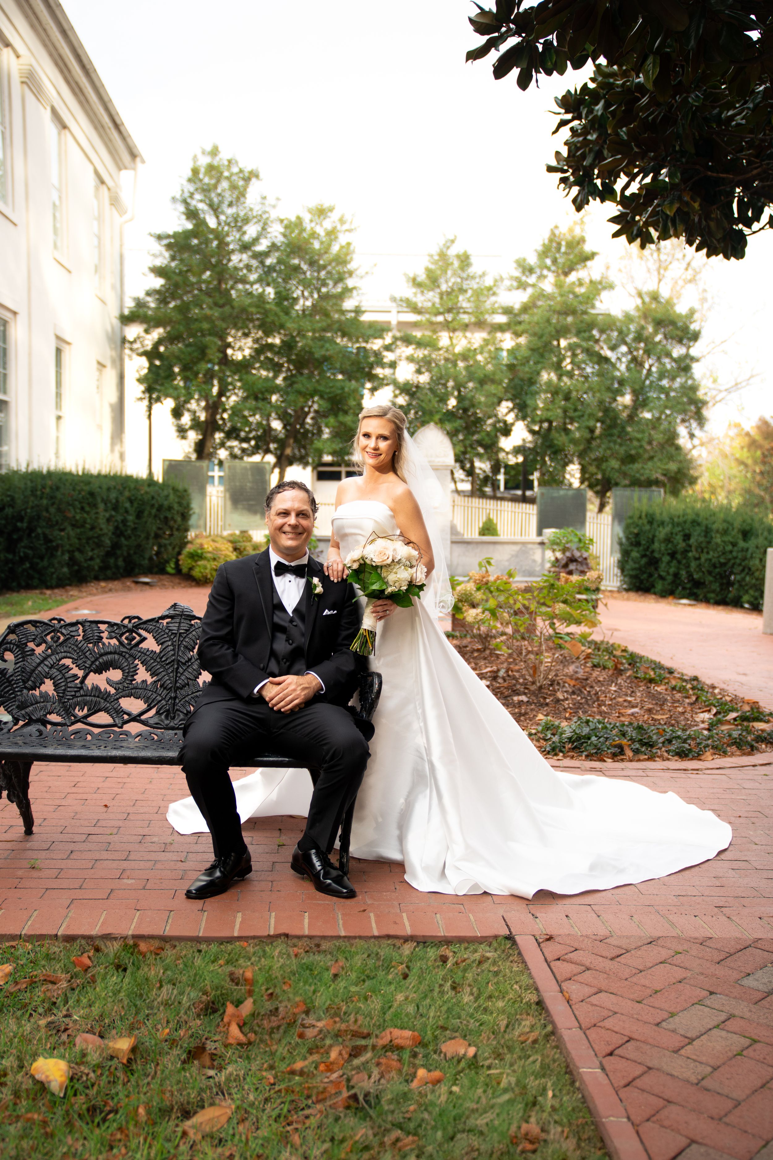 A wedding couple sits on brick pavement surrounded by autumn leaves and white columns.