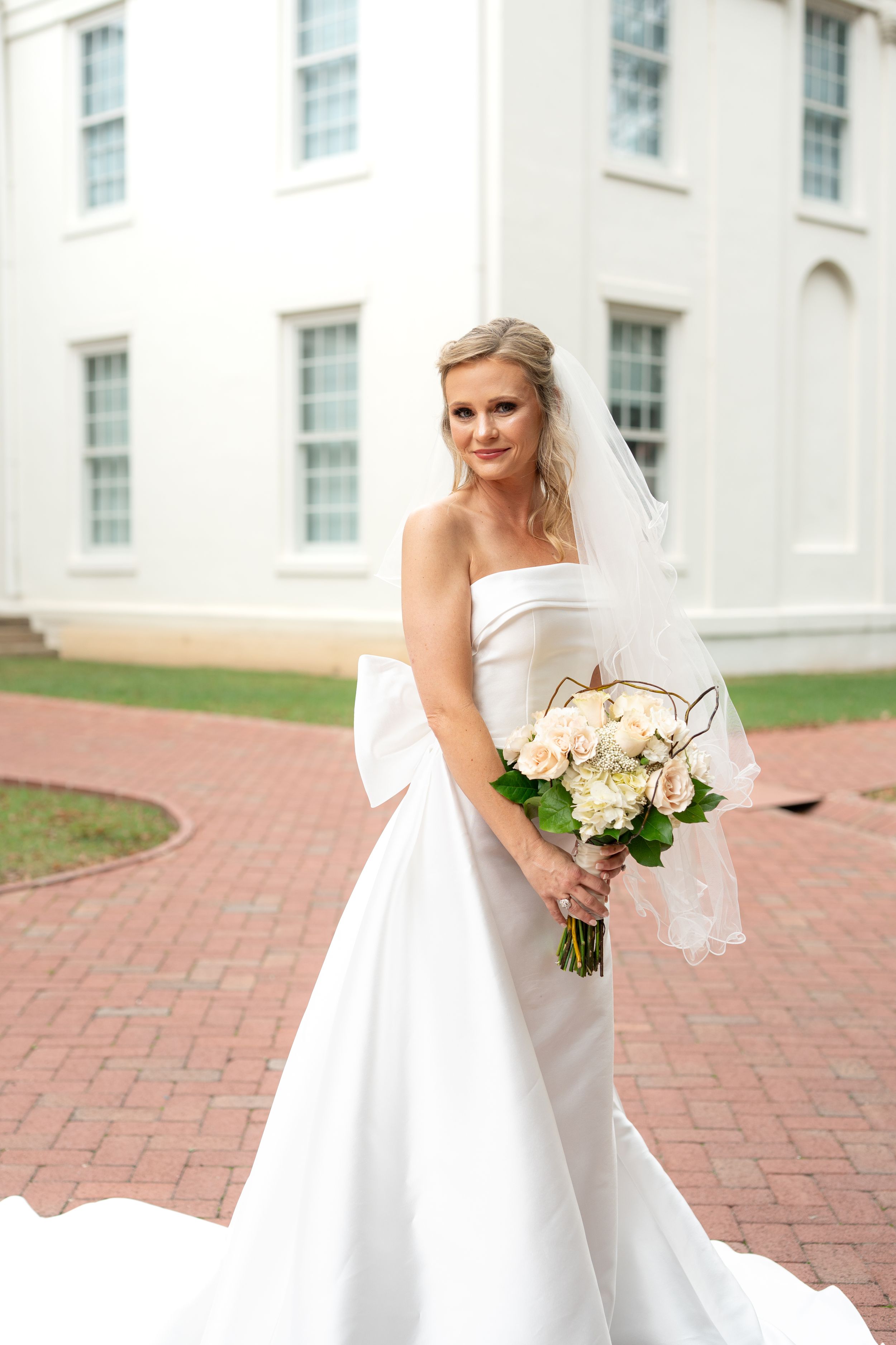 Elegant bride in white gown holds bouquet on brick courtyard.