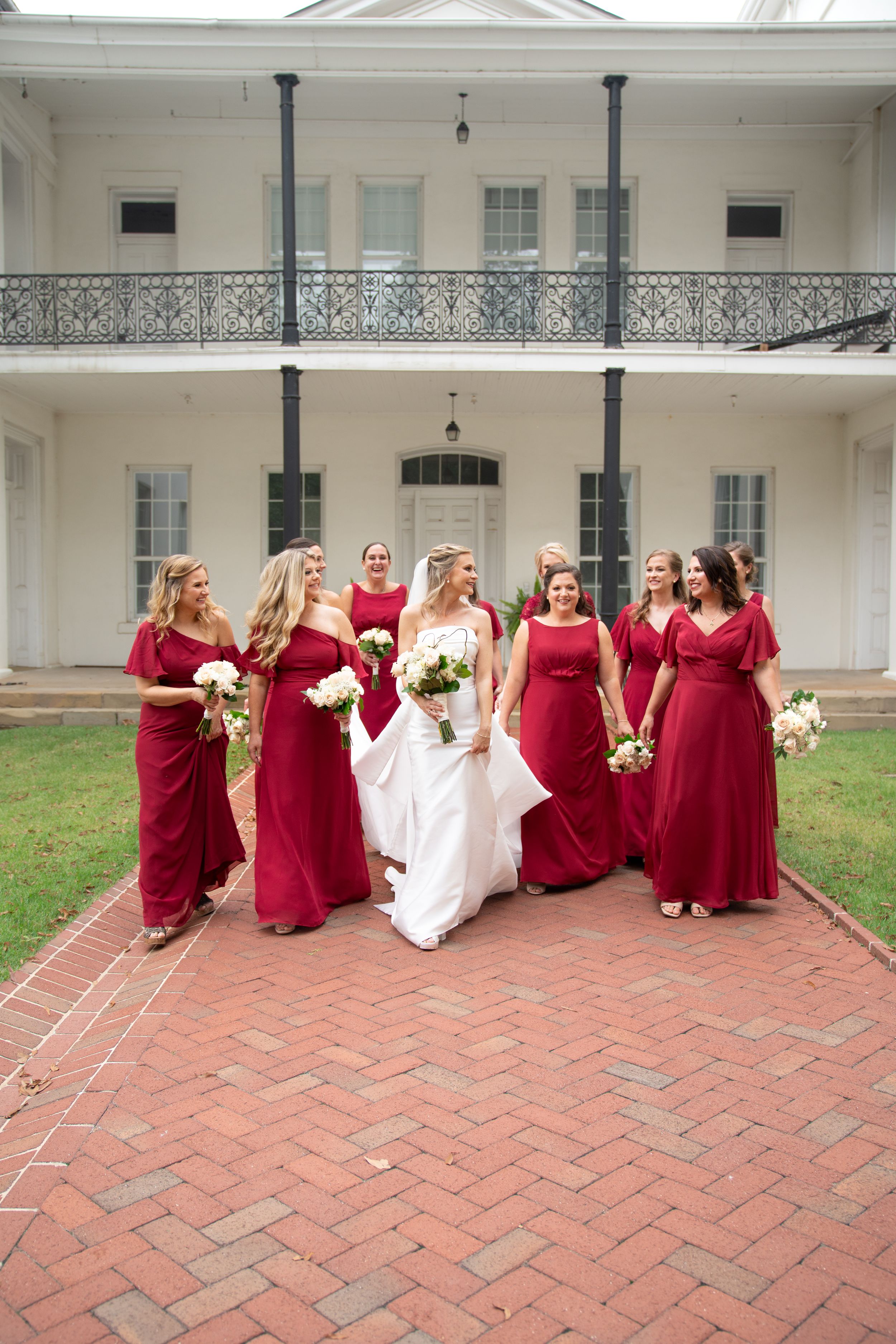 Wedding party in red and white dresses poses on brick patio in front of white building.