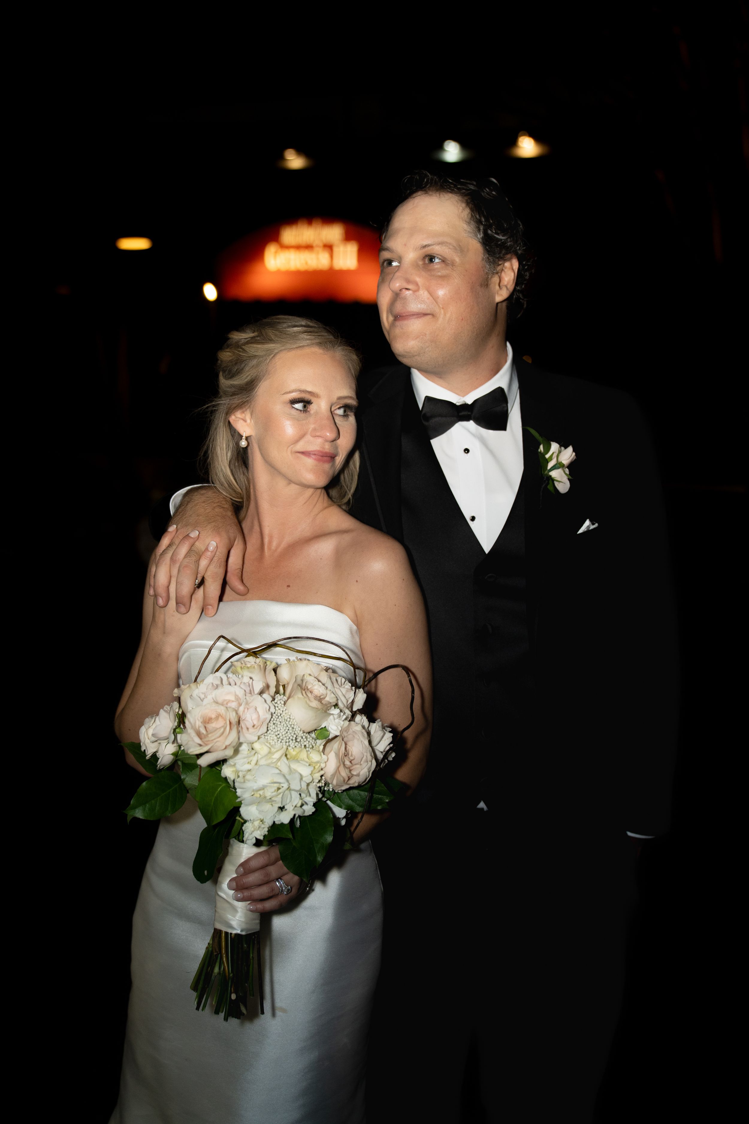 A bride in white holds a bouquet while dancing with her groom in formal attire at an evening reception.