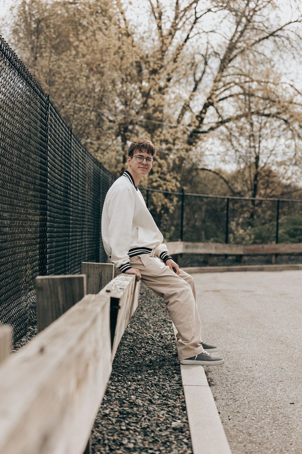 Person in white sweater and beige pants sits casually on concrete ledge by chain link fence on autumn day.