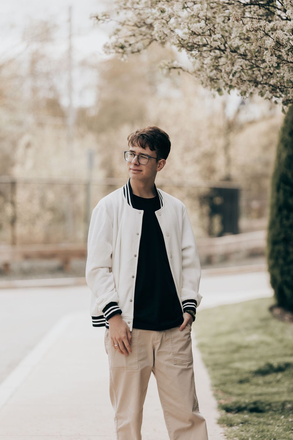 Young person wearing a white varsity jacket with black trim, glasses and khaki pants stands on a path near trees.