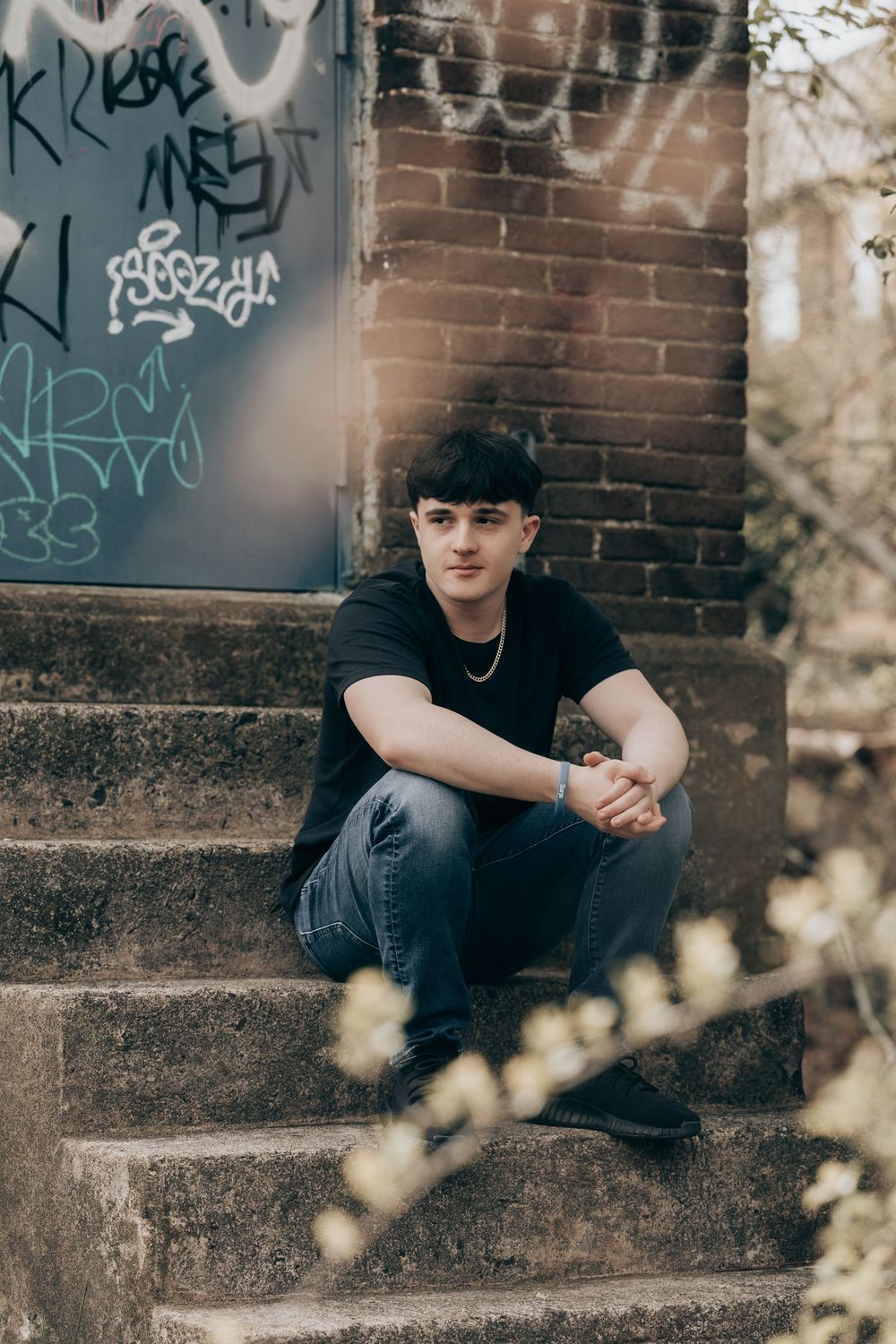 A person sits on concrete steps near a brick wall with graffiti, wearing black clothing in a moody outdoor setting.