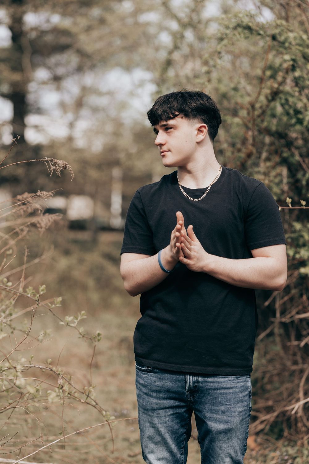 A person wearing a black t-shirt and jeans stands outdoors with hands pressed together in a reflective pose.
