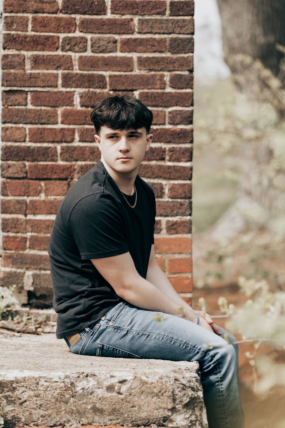Person in black t-shirt and jeans sits casually against brick wall in outdoor portrait setting.