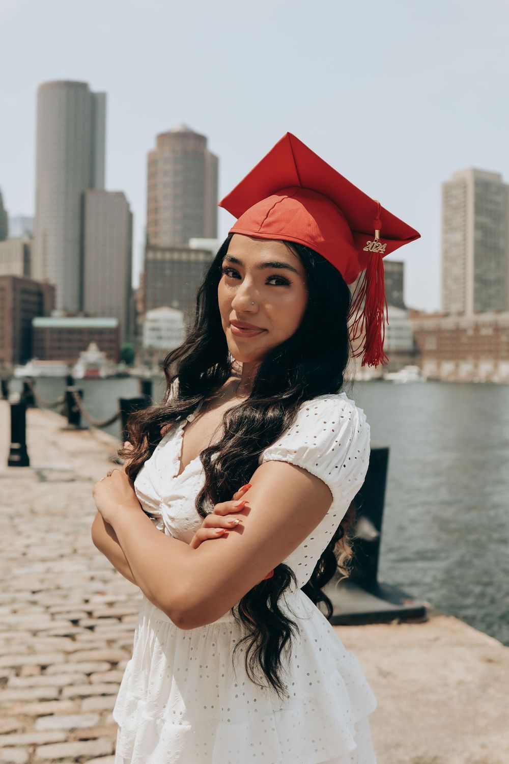 Graduate in white dress and red cap posing with Boston skyline background.