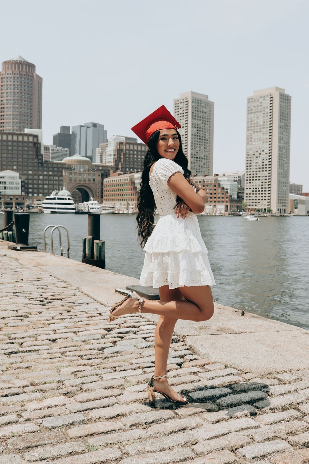 Graduate in white dress strikes playful pose on cobblestone pier with city skyline behind.