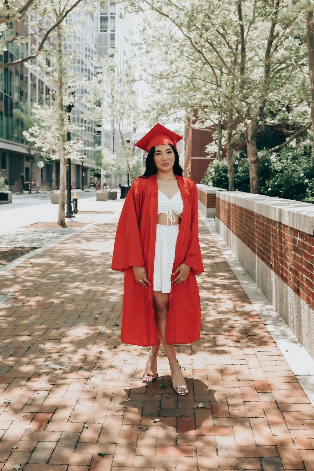 Graduate in red cap and gown poses on brick walkway with blooming trees in urban setting.