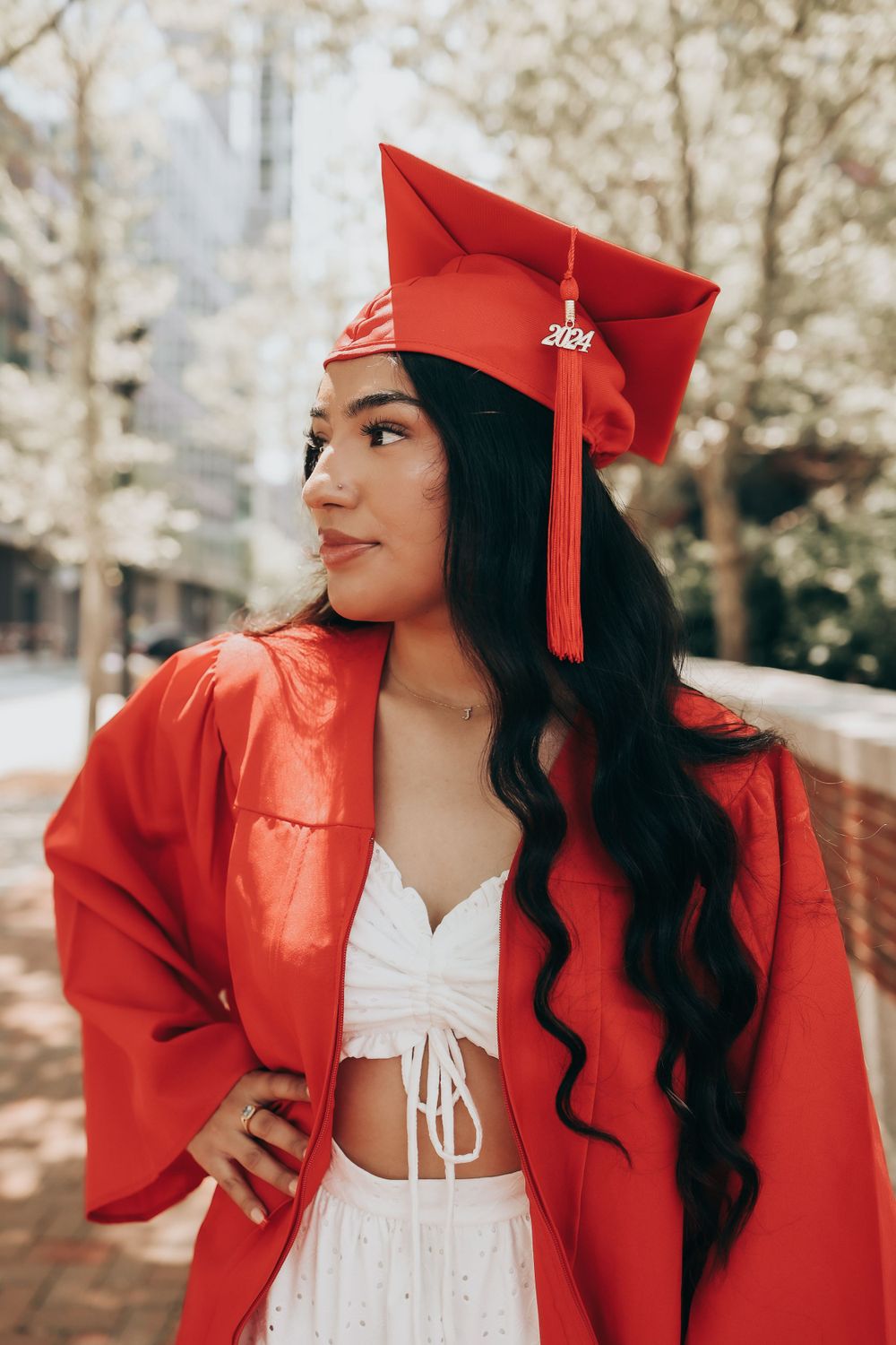 Graduate in red academic regalia with flowing dark hair poses against brick wall outdoors.