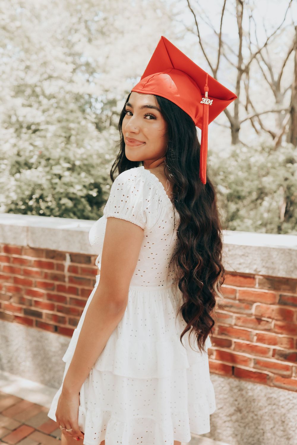 Graduate wearing white dress and red cap poses by brick wall with spring trees in background.