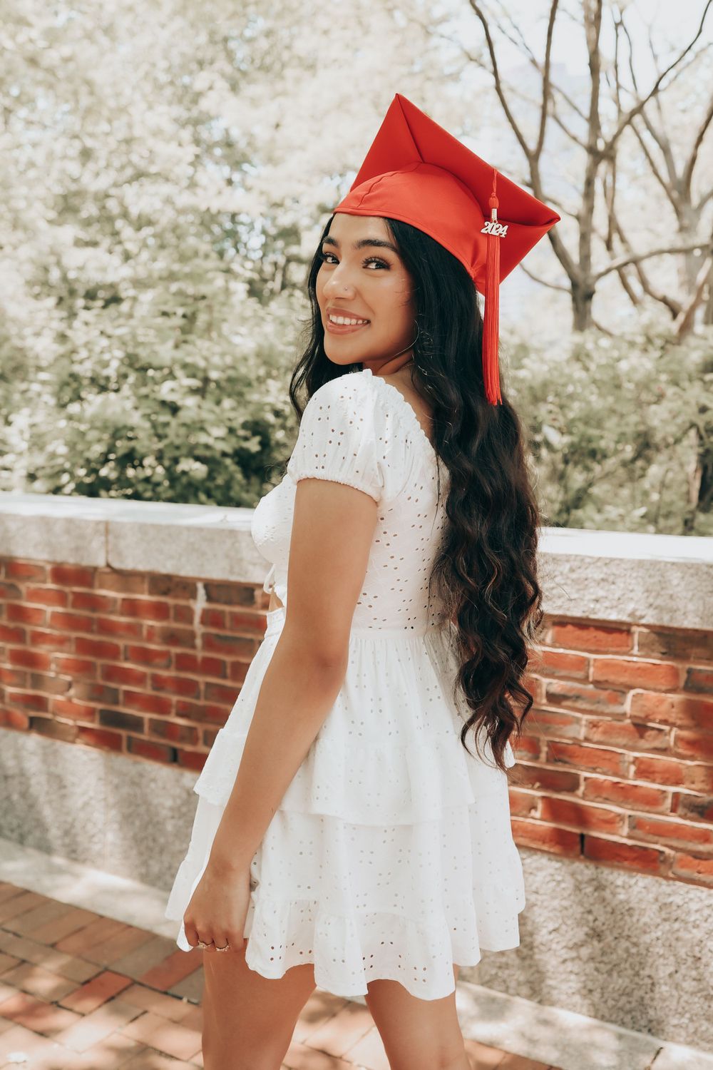 Graduate in white eyelet dress and red cap poses on brick pathway with cheerful expression.