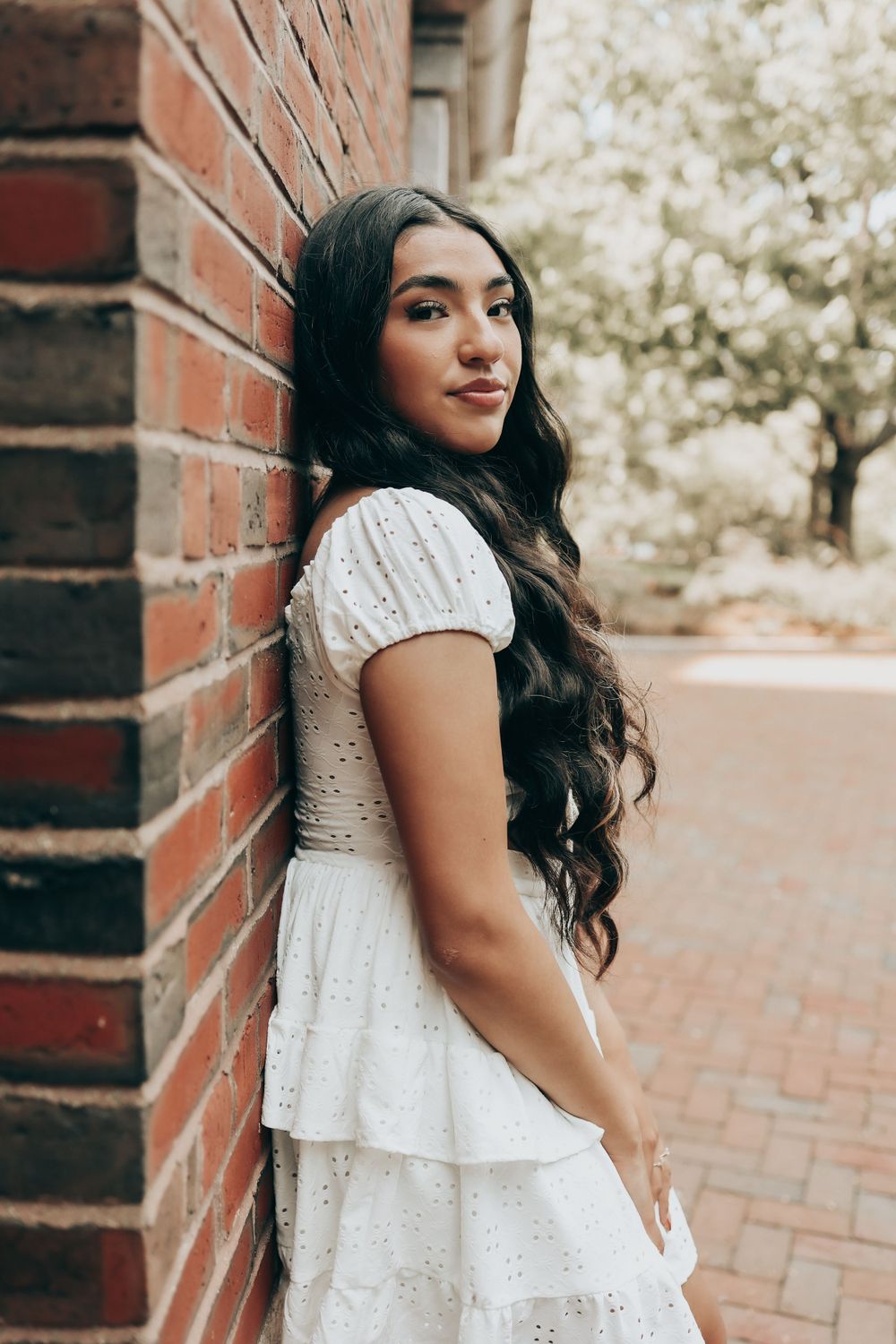 Graduate in white lace dress poses against red brick wall on sunny spring day.
